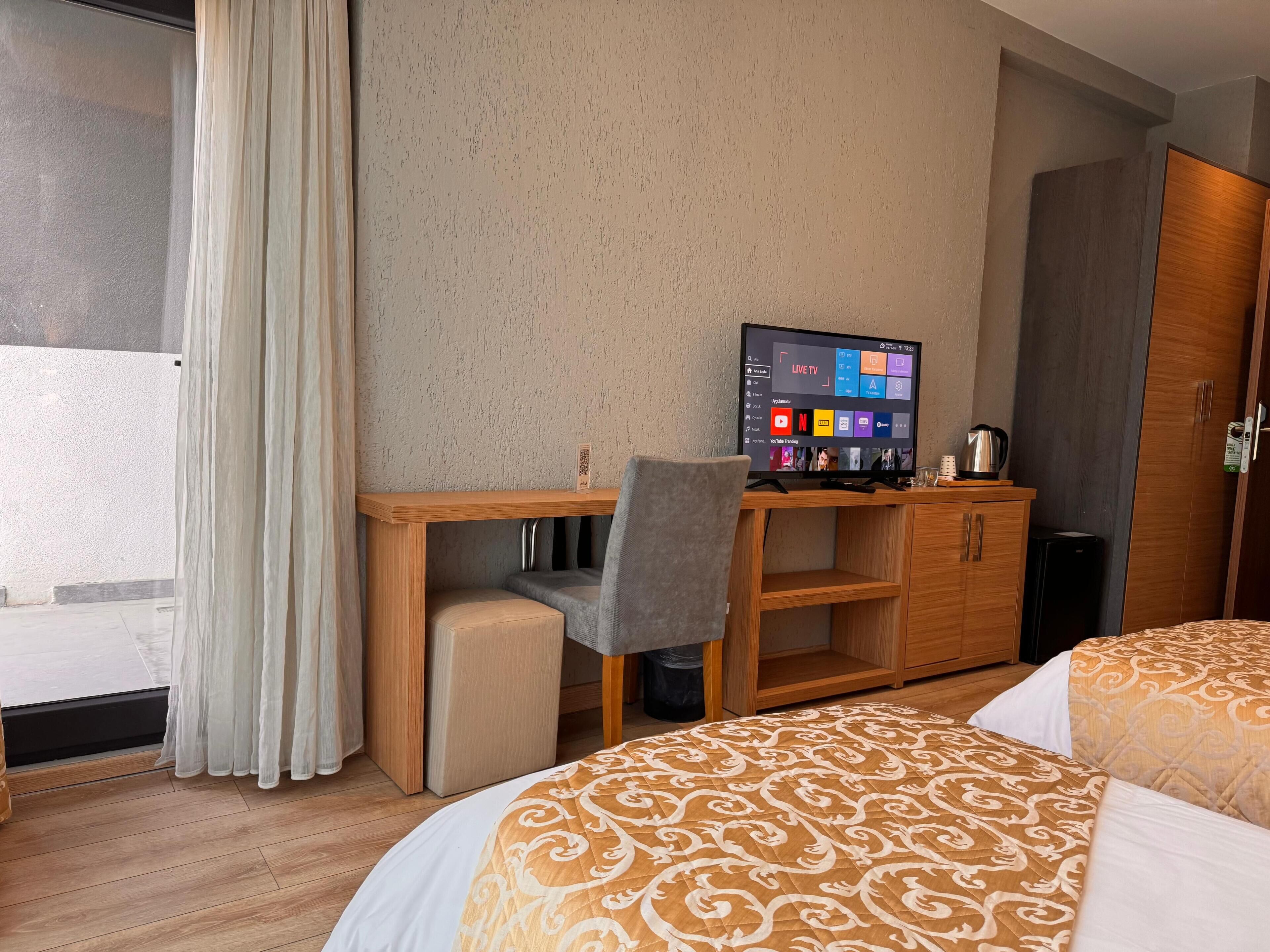 Quarto de hotel com cama, escrivaninha com cadeira, televisão e guarda-roupa.