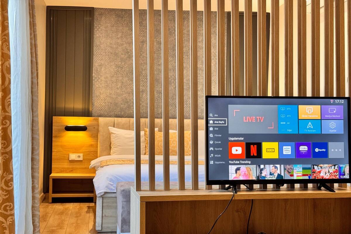 A hotel room with a bed, a wooden TV stand, and a TV displaying a live TV channel interface.