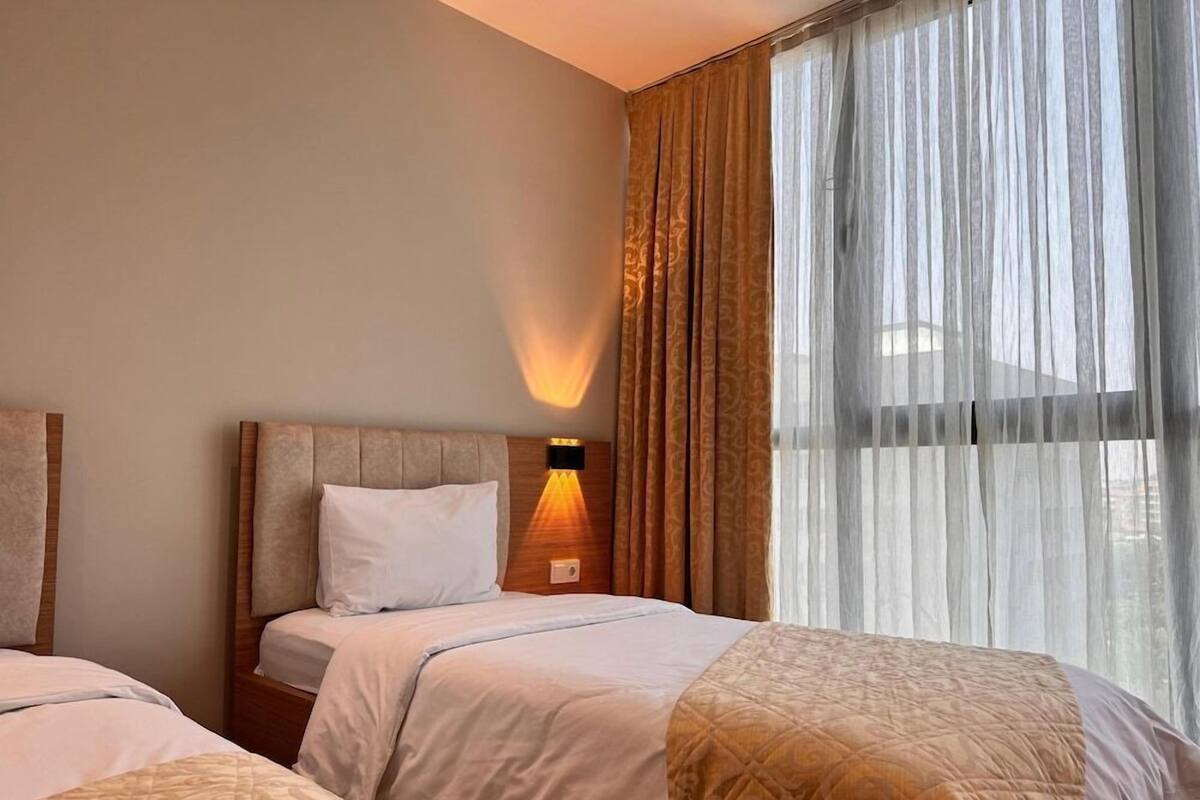 A hotel room with two beds, a headboard, a wall-mounted lamp, and a window with sheer curtains.