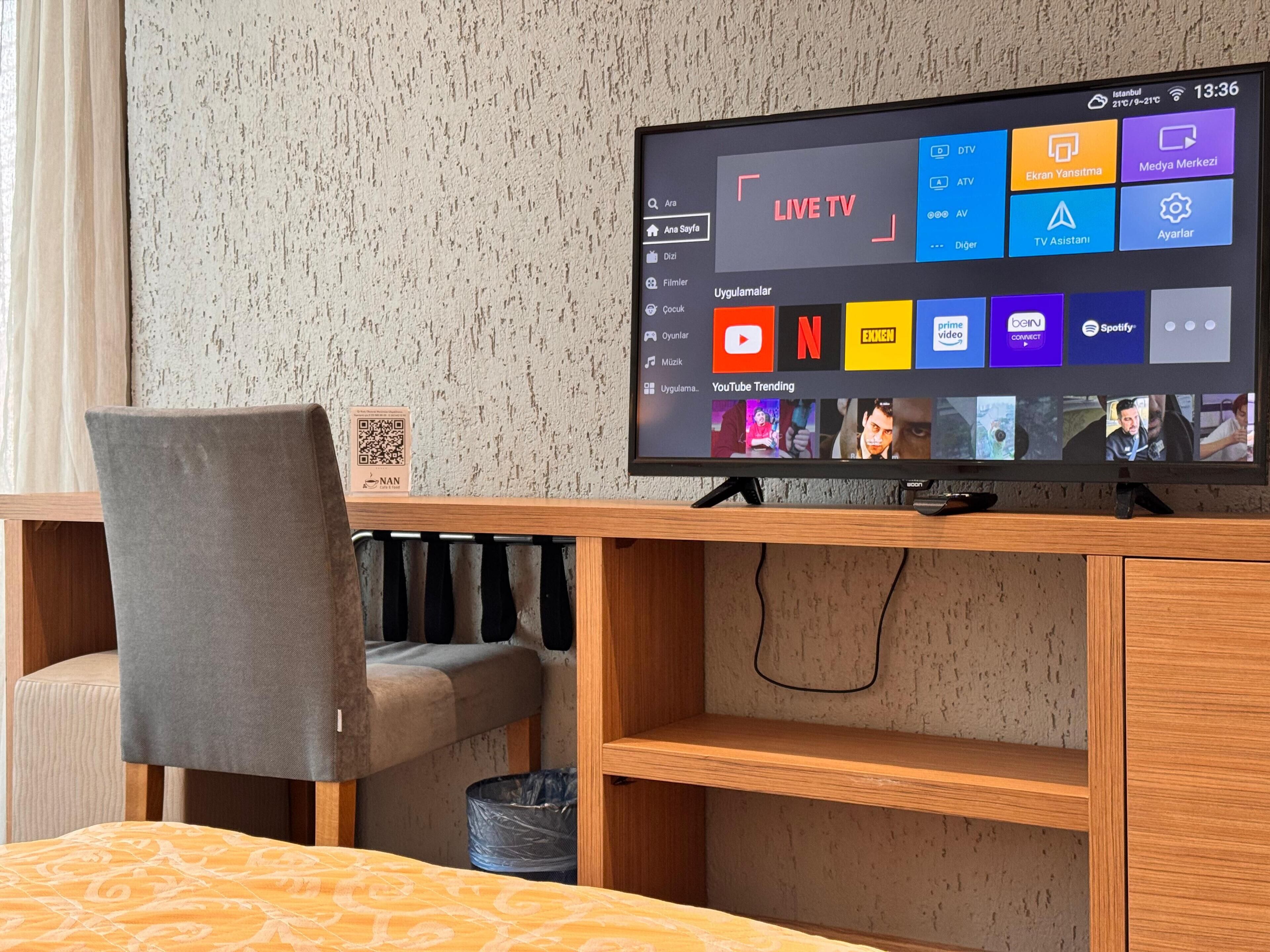 A hotel room with a flat-screen TV displaying a live TV channel, a wooden desk, a grey chair, and a bed with a patterned bedspread.