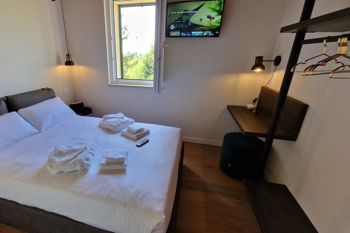 A neatly made bed with white linens and towels, a television mounted on the wall, and a small table with a lamp and a coat rack.