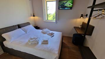 A neatly made bed with white linens and towels, a television mounted on the wall, and a small table with a lamp and a coat rack.