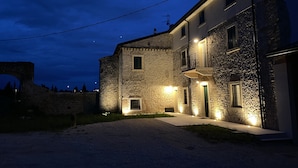 A stone building with a balcony, illuminated by exterior lights, and a stone archway in the background. - CasaOssan (San Pietro in Cariano)