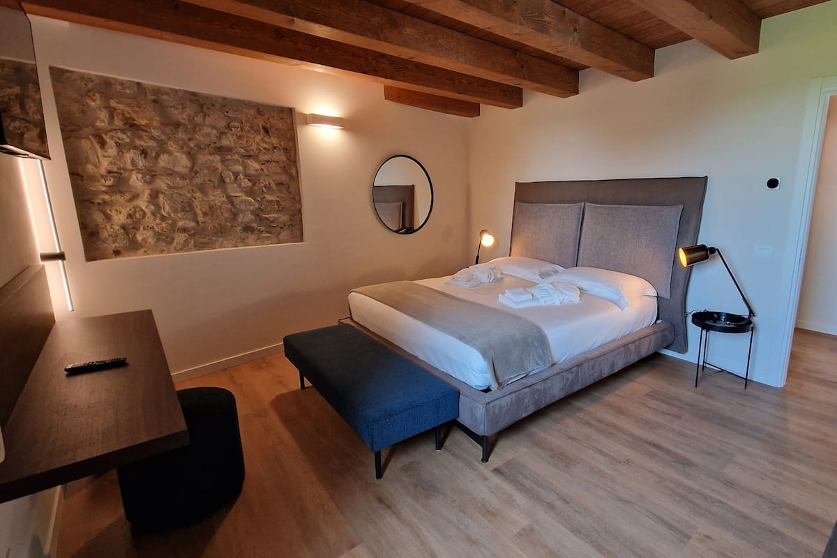 A bedroom with a bed, bedside table, bench, lamp, and a stone wall.