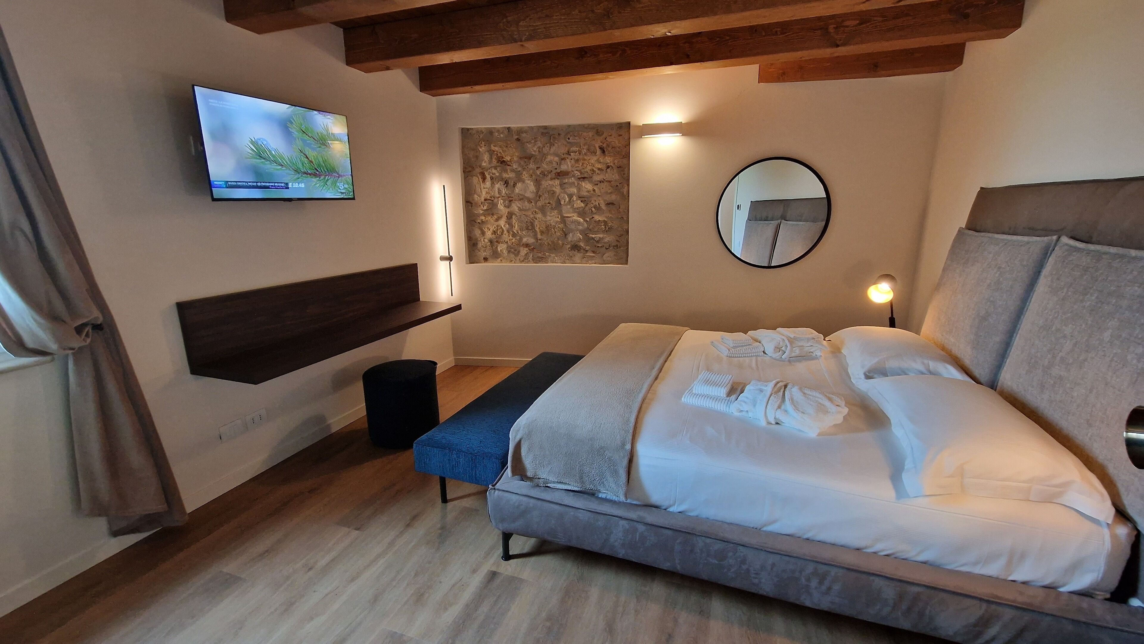 Quarto moderno com cama, TV montada, painel decorativo de pedra, espelho redondo e teto de madeira.