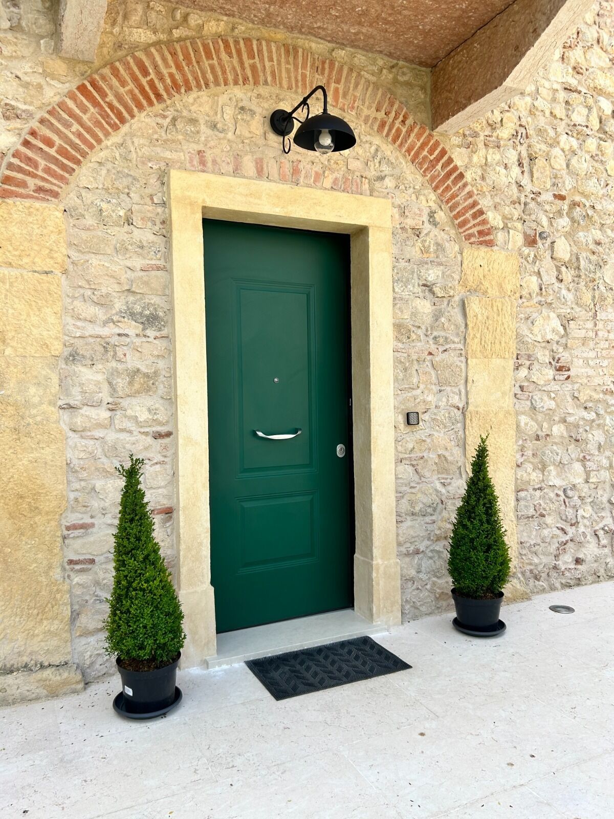 A green door with a handle, flanked by two potted evergreen shrubs, set in a stone building with a brick archway above.