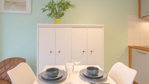 Uma área de refeições com uma mesa branca posta para dois, uma planta em vaso na parede e um armário branco com portas.