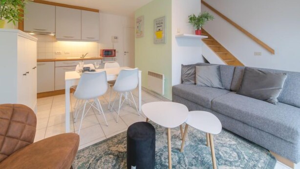 Uma sala de estar moderna com um sofá cinzento, uma poltrona castanha e uma área de jantar com cadeiras brancas e uma mesa pequena.