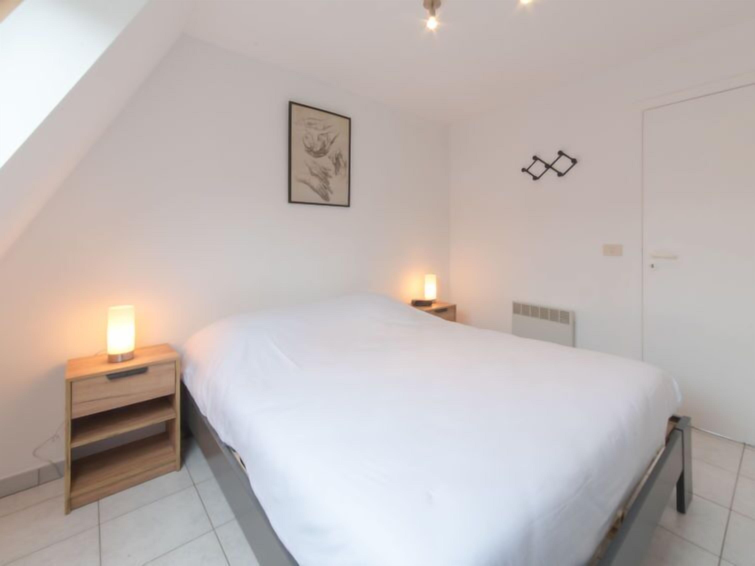 Ein Schlafzimmer mit einem Bett, zwei Nachttischen inklusive Lampen, einer weißen Wand mit einem gerahmten Bild, einer Tür und einer geneigten Decke.