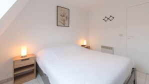 Quarto com cama, duas mesas de cabeceira com luminárias, parede branca com quadro emoldurado, porta e teto inclinado.