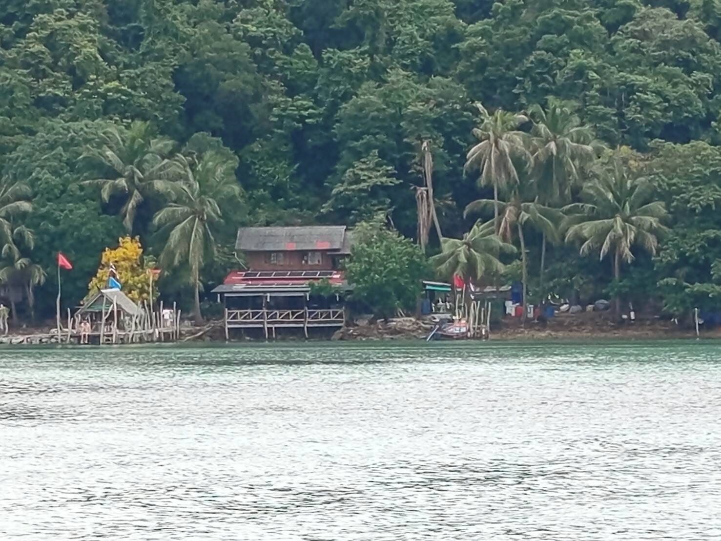 A house with a red roof and a small porch, surrounded by palm trees and a body of water.