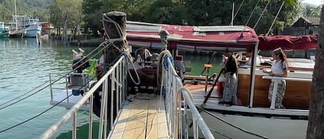 Un muelle de madera con barandilla metálica, barcos amarrados al fondo y una persona de pie sobre el muelle.
