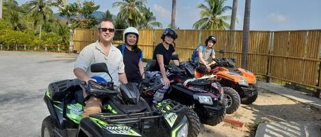 Three people on ATVs parked in a sandy area with a wooden fence and palm trees in the background.