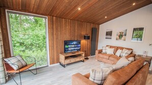 Un salón moderno con techo de madera, un televisor grande sobre un mueble de madera, un sofá marrón y una ventana con persianas.