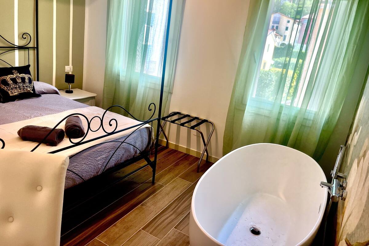 A bedroom with a four-poster bed, a large white bathtub, and a view of the outside through sheer curtains.
