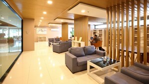 A modern lobby with a glass wall, grey sofas, a coffee table, wooden partitions, and a person working at a desk. - Vacation Home Rental The Starhill Condo Chiang Mai (Muang Chiangmai)