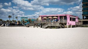 A beachside bar with a wooden deck and a pink facade, located near a parking area and a building with a sign that reads "PINK PONY PUB EST. 1956".