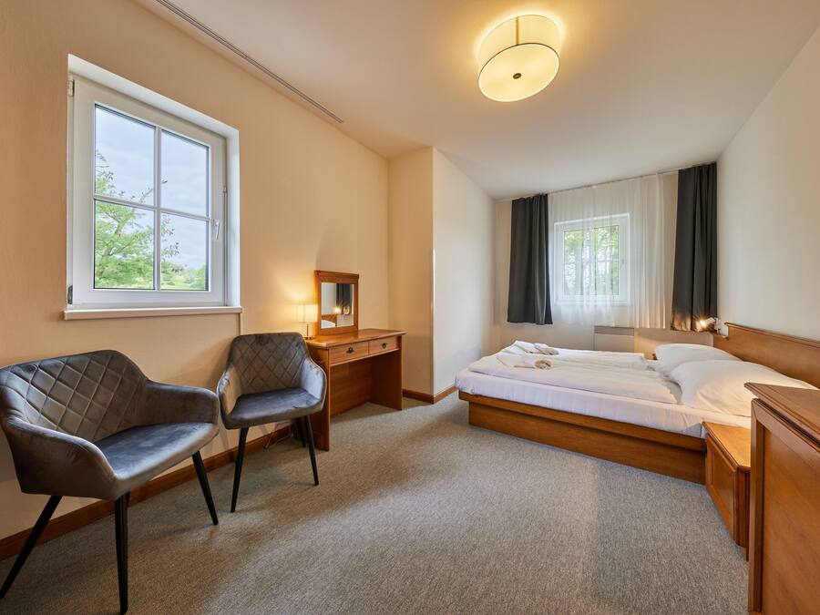 A hotel room with a bed, a desk, two chairs, a mirror, and a window with curtains.