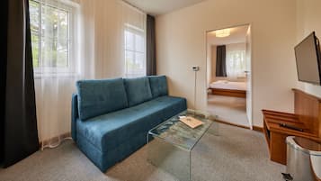 A room with a blue sofa, a glass coffee table, a wooden side table, a bed, and a TV.