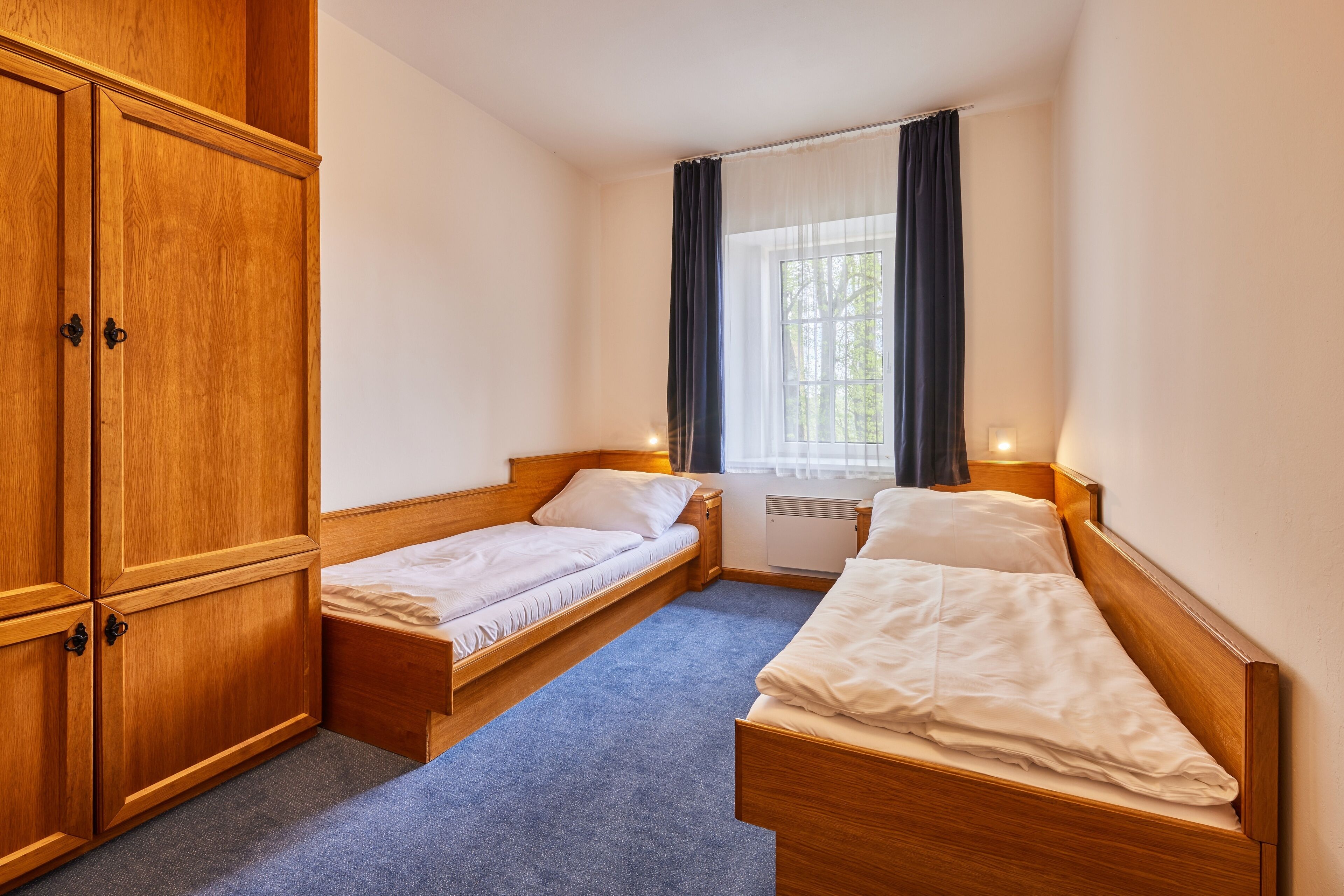 A small, well-lit room with two single beds, a wooden wardrobe, and a window with curtains.