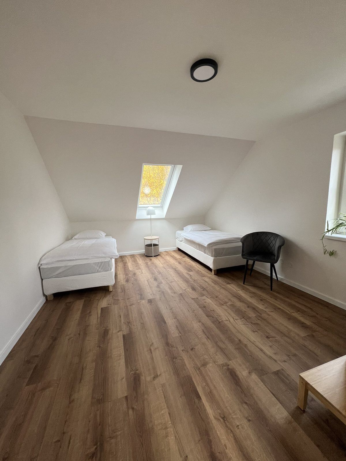 Un dormitorio pequeño con techo inclinado, dos camas, una silla y una mesita.