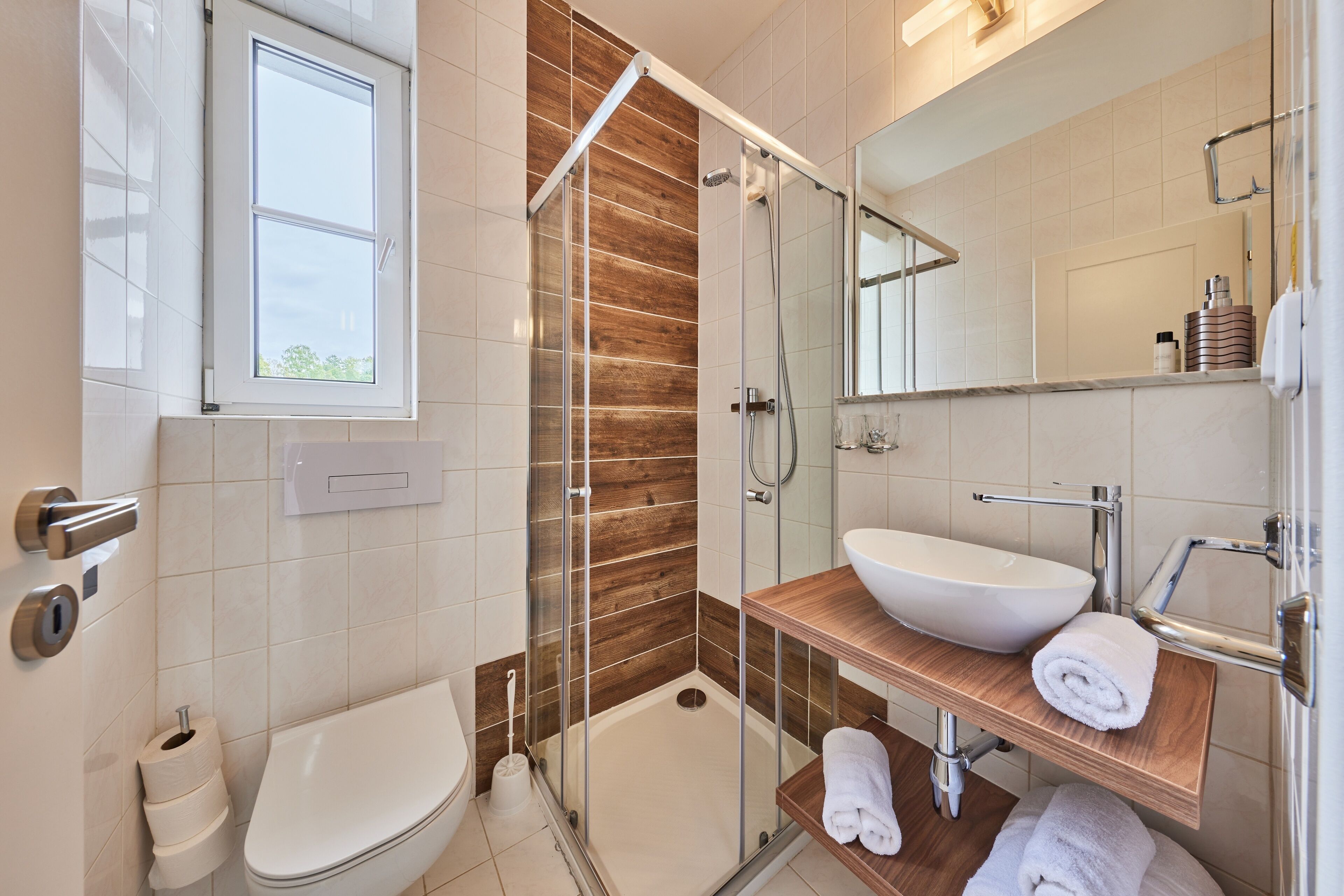 Un baño moderno con ducha envuelta en vidrio, inodoro blanco y tocador de madera con lavamanos y toallero.