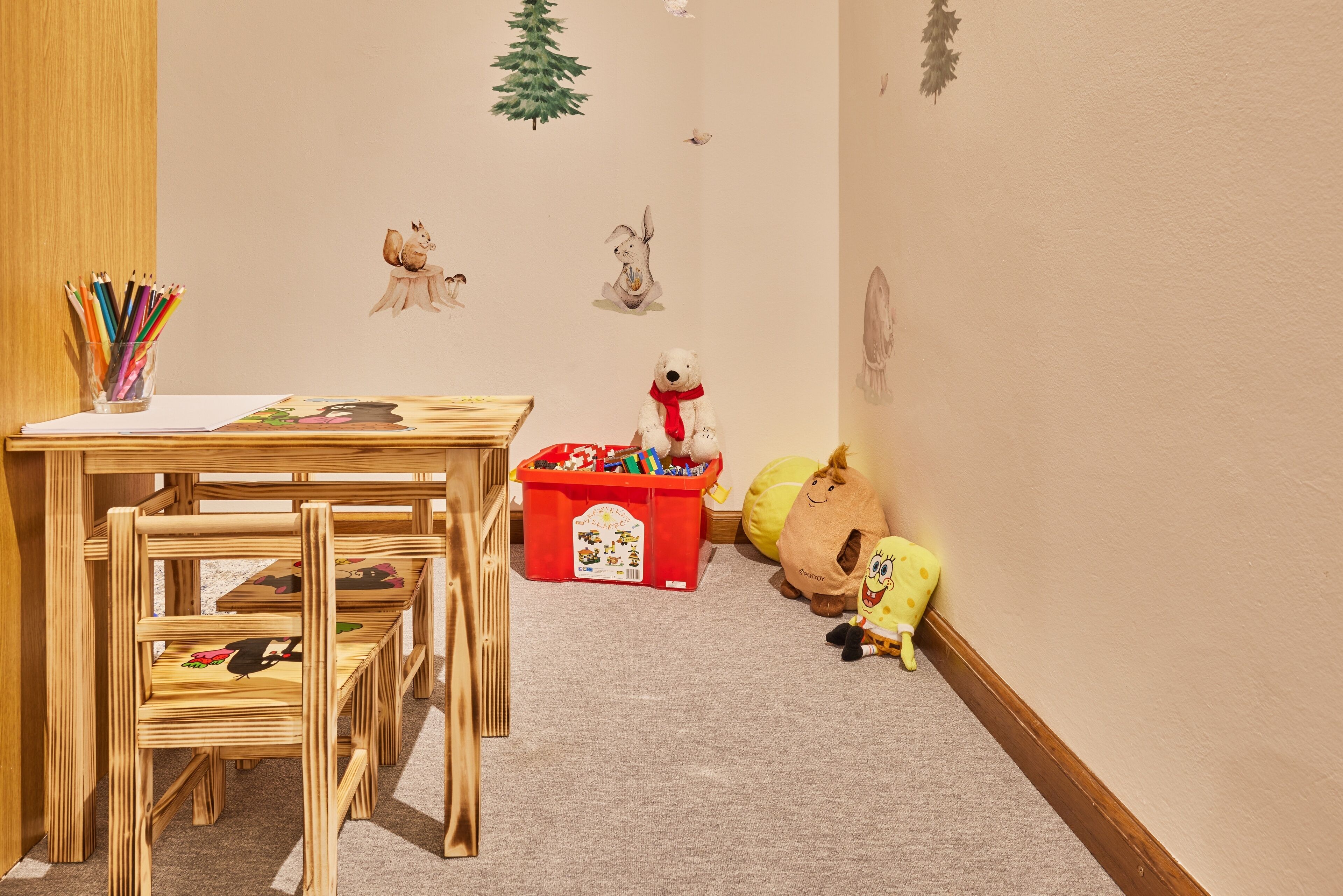 A children's playroom with wooden chairs, a table with art supplies, a red storage bin, and wall decorations including a bear and a tree.