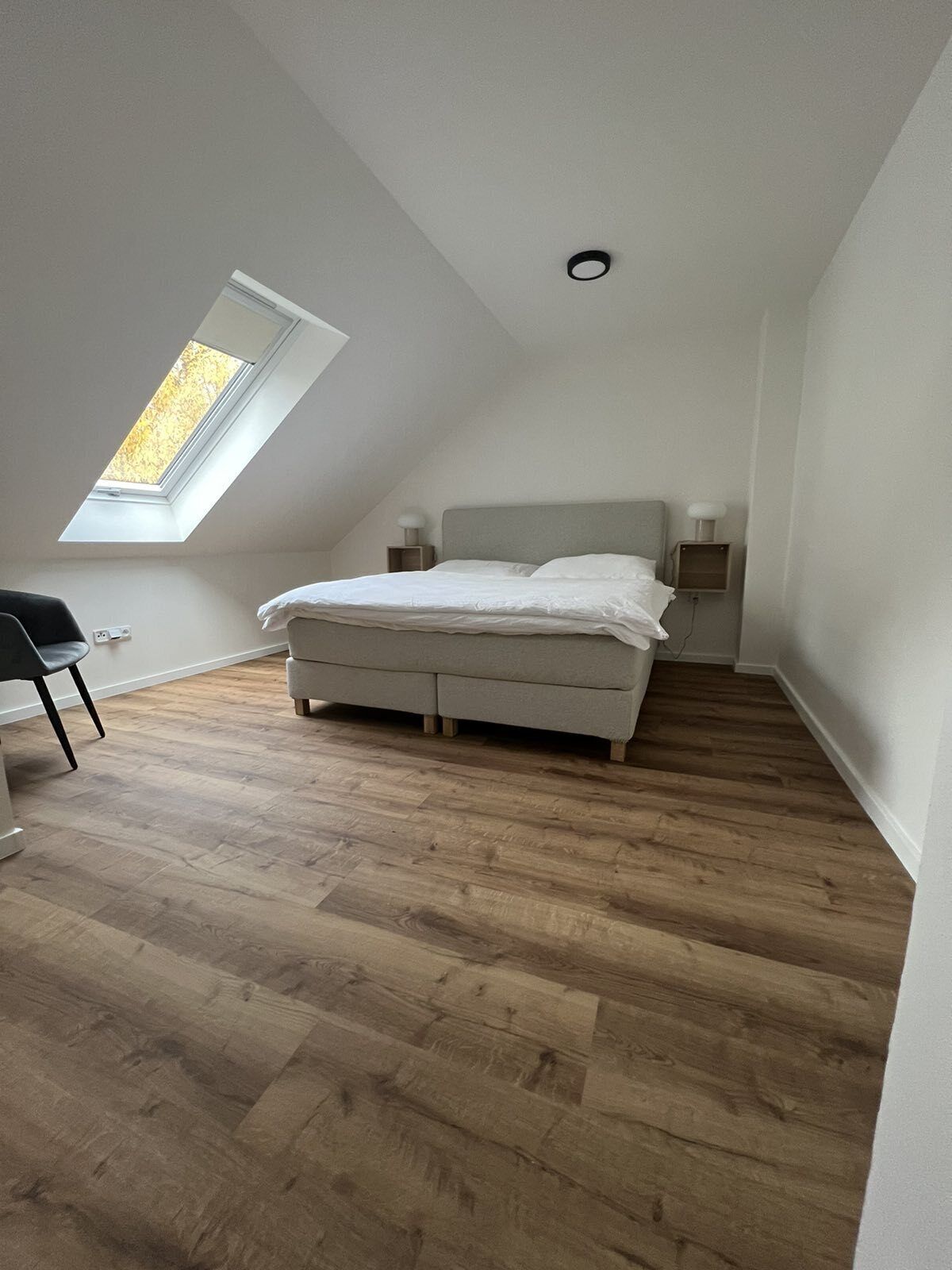 Un dormitorio con techo inclinado, tragaluz, cama, lámparas de noche y una silla.