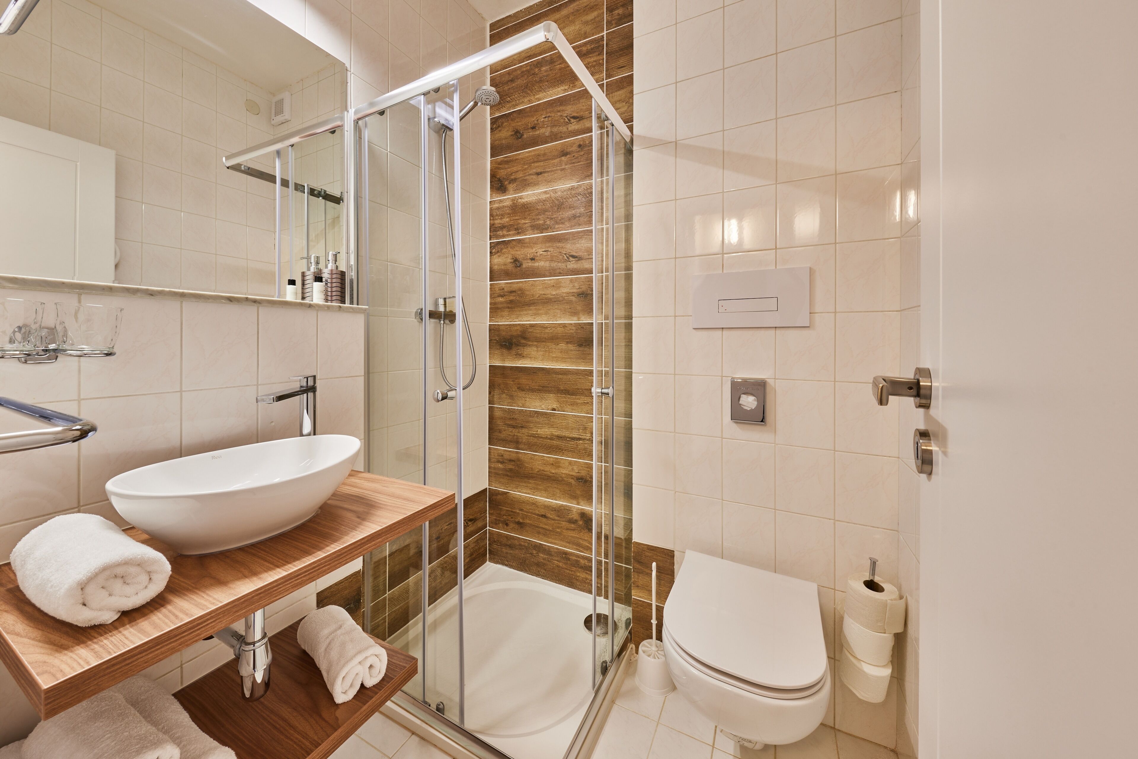 Un baño moderno con ducha de vidrio, tocador de madera, lavamanos blanco y retrete.