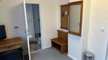 A bedroom with a wooden desk, a chair, a wardrobe, a mirror, a trash bin, and a shower area.