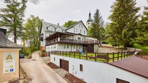 A large, white building with a wooden balcony and a blue slide, surrounded by trees and a small parking area.