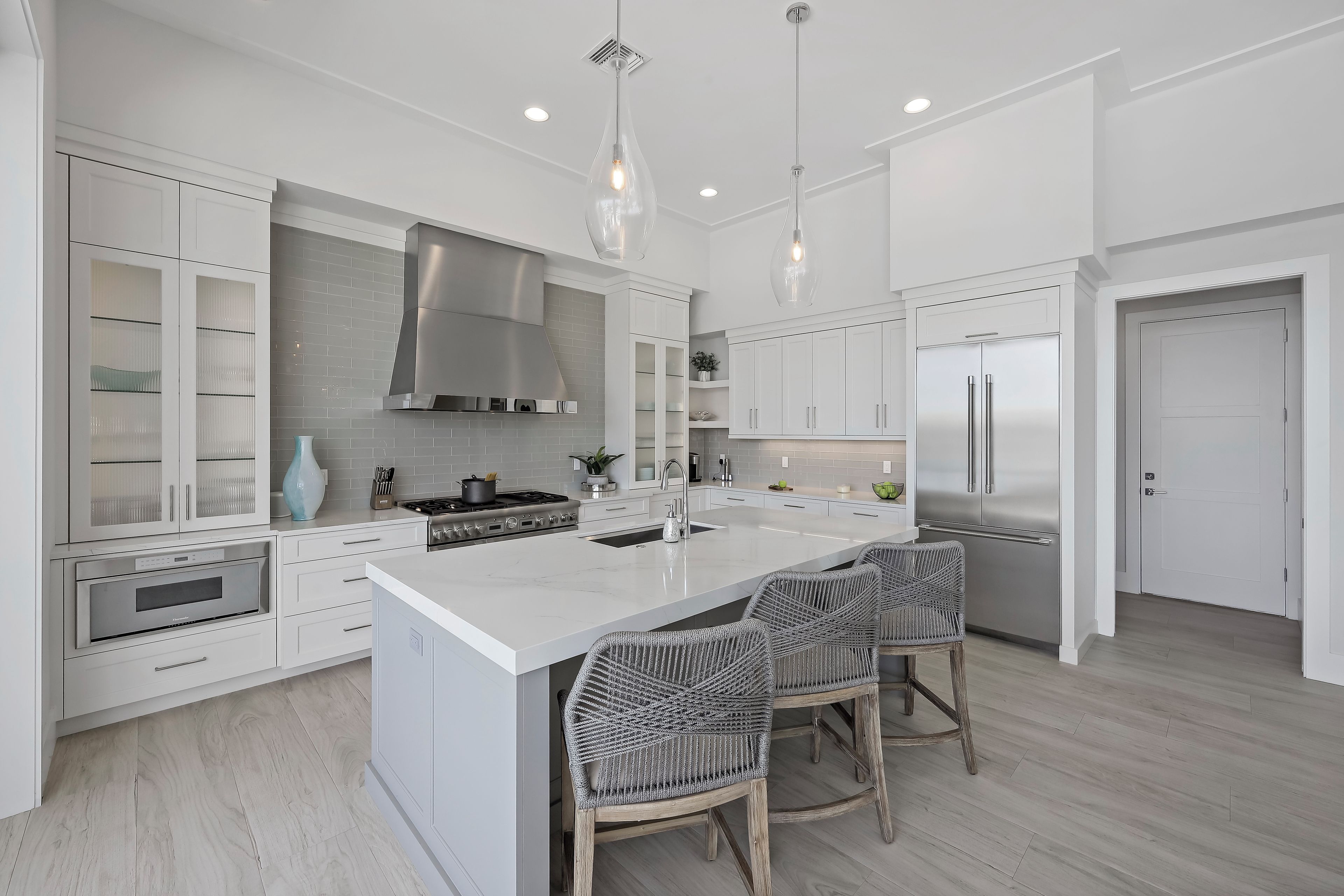 A modern kitchen with white cabinetry, a central island, stainless steel appliances, and wicker chairs.