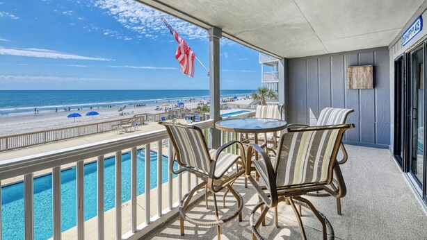 A balcony with a view of the beach, a pool, and a flag.