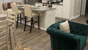 A modern kitchen with white cabinets, a stainless steel refrigerator, and a microwave. There is a dining area with a table and chairs, and a teal sofa in the living room.