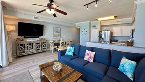 A living room with a sofa, coffee table, TV, dining area, kitchen, ceiling fan, and clock. - Gulf Front 3rd Floor Perfect Location Free Beach Chairs! Free Golf Everyday! (Panama City Beach)