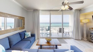 A room with a blue sofa, a wooden coffee table, a ceiling fan, and a view of the beach. - Gulf Front 3rd Floor Perfect Location Free Beach Chairs! Free Golf Everyday! (Panama City Beach)