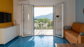 Et værelse med blå flisegulv, en hvid sofa, et fjernsyn på et hvidt skab, og en balkon med et bord og stole.