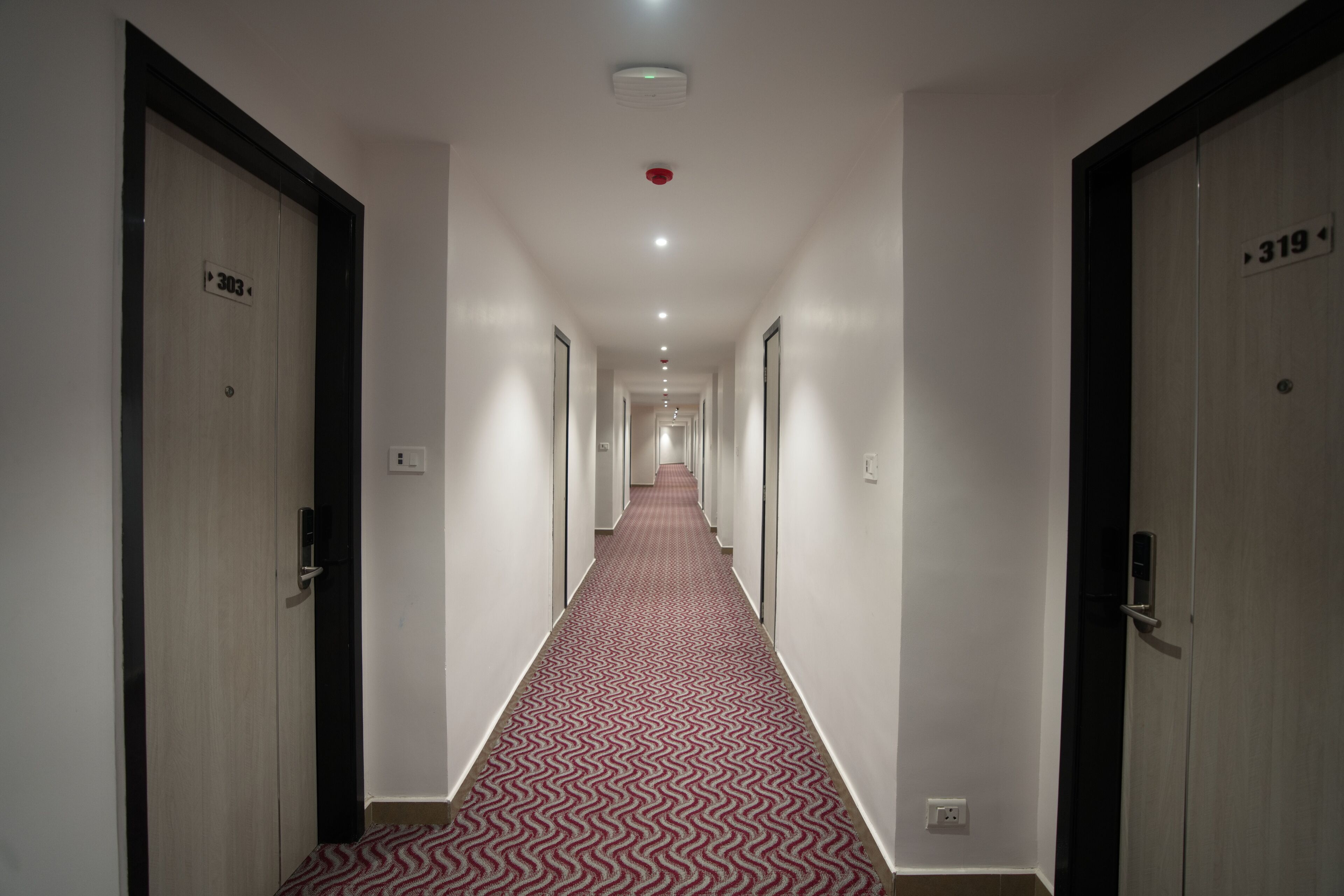 A hotel hallway with a patterned carpet, doors numbered 303 and 319, and a red emergency light on the ceiling.
