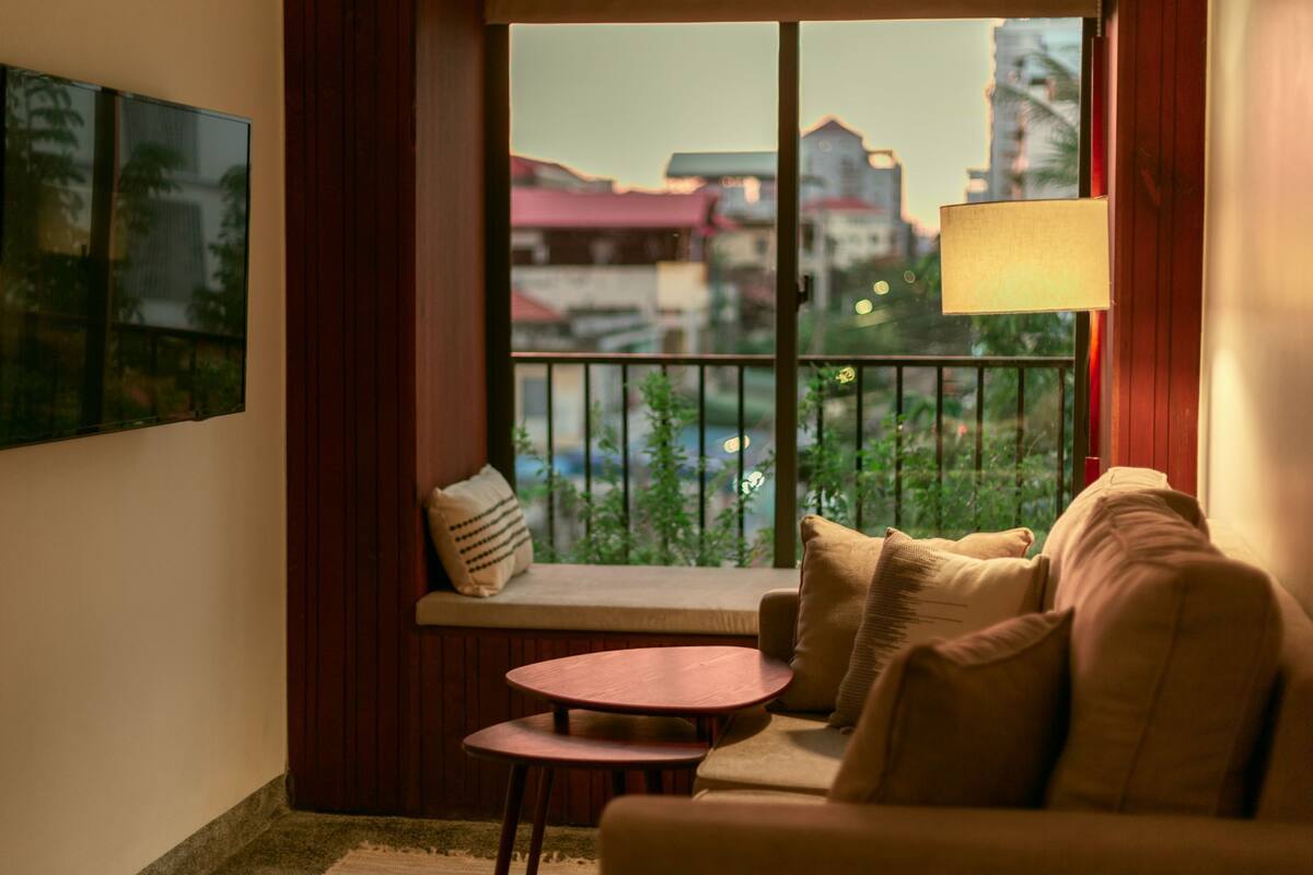 Um quarto aconchegante com sofá, uma mesinha redonda e janela com vista para edifícios e áreas verdes.
