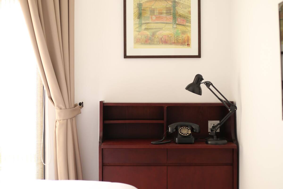 Quarto com cama, escrivaninha com luminária, telefone e quadro na parede.