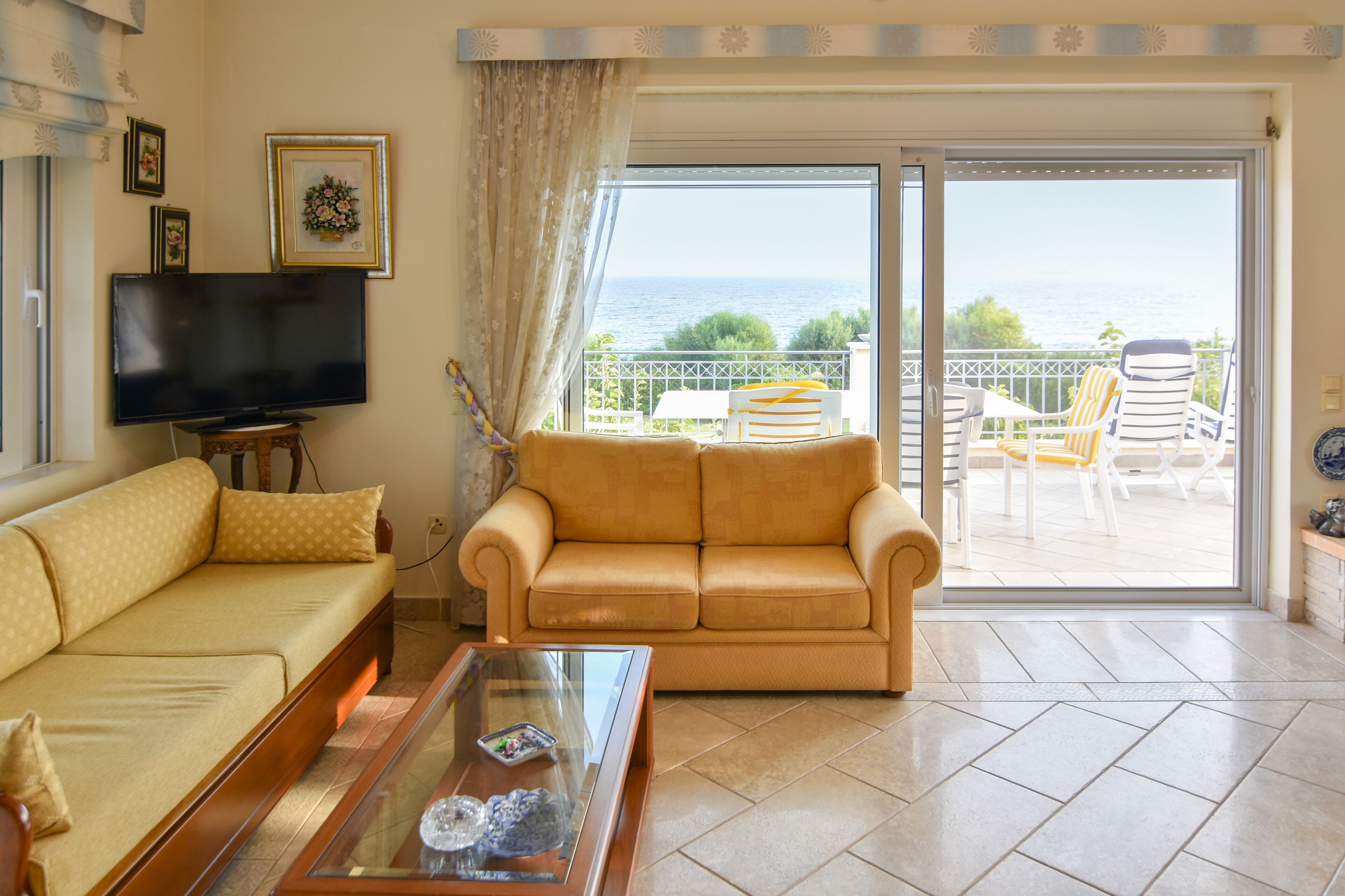 A living room with a sofa, a coffee table, a TV, and a balcony with chairs and a view of the sea.