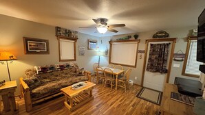 Una sala de estar acogedora con piso de madera, un sofá, una mesa de centro, una mesa de comedor con sillas, un ventilador de techo y diversas decoraciones en las paredes.