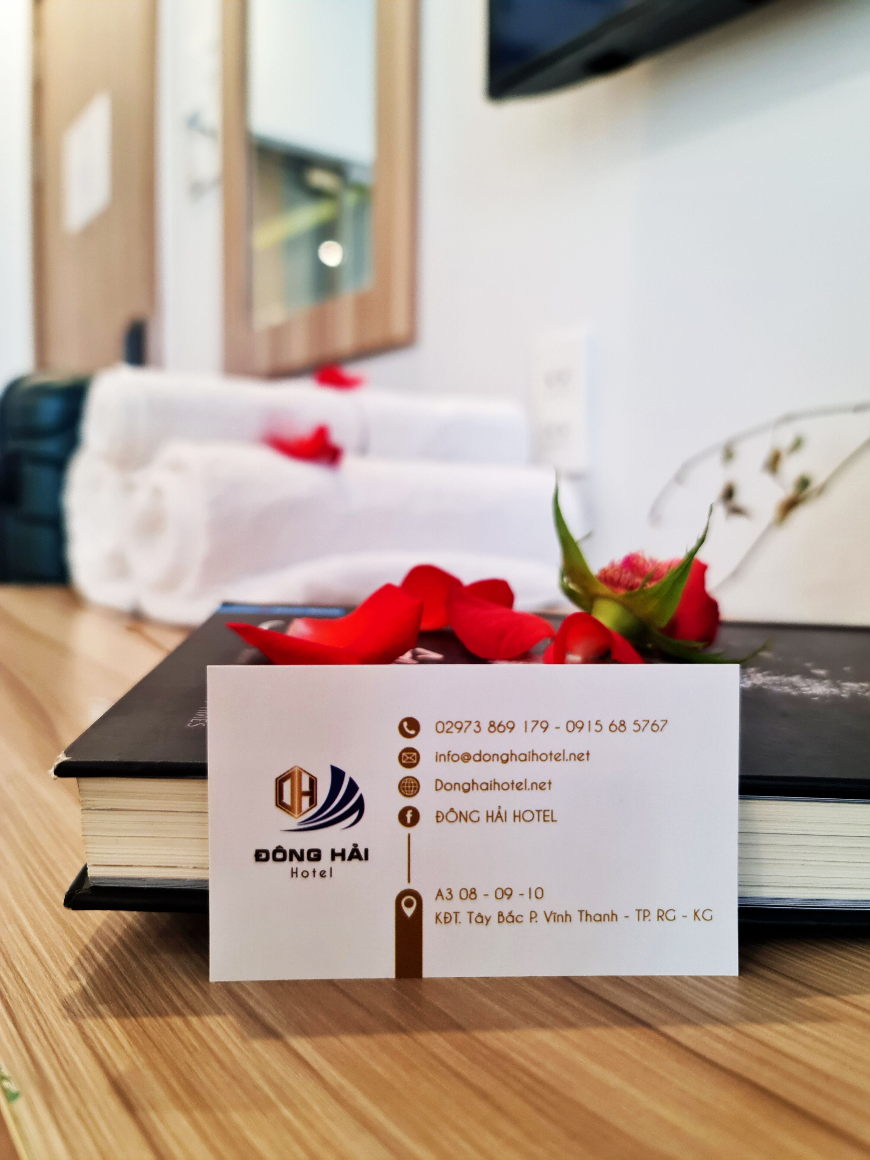 A hotel business card with contact information and the hotel's name, Dong Hai Hotel, placed on a wooden table with a book and a bouquet of red flowers.