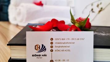 Une carte de visite d’hôtel indiquant les coordonnées et le nom de l’hôtel, « Dong Hai Hotel », est posée sur une table en bois, à côté d’un livre et d’un bouquet de fleurs rouges.