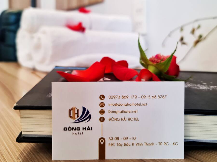 Une carte de visite d’hôtel indiquant les coordonnées et le nom de l’hôtel, « Dong Hai Hotel », est posée sur une table en bois, à côté d’un livre et d’un bouquet de fleurs rouges.