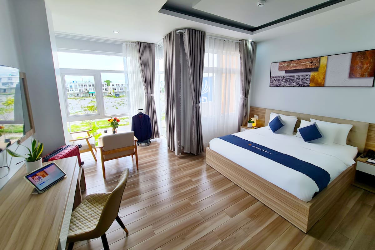 A modern hotel room with a large bed, a desk with a chair, a small table with a potted plant, and a view of the outside.