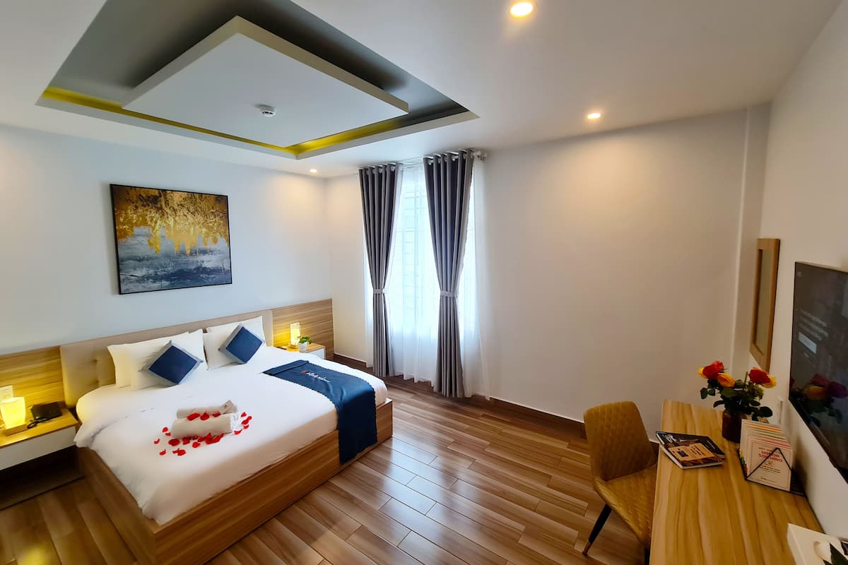 A modern hotel room with a large bed, a bedside table with a lamp, a desk with a chair, and a large window with curtains.