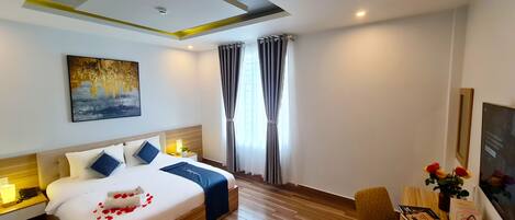 Una habitación de hotel moderna con una cama grande, una mesita de noche con lámpara, un escritorio con silla y un amplio ventanal con cortinas.