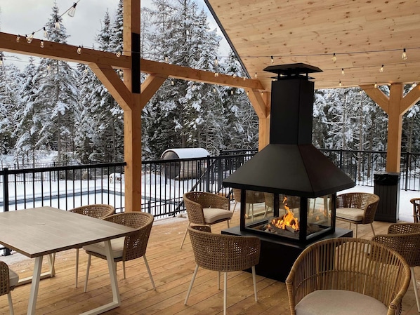 Ett takförsett utomhusområde med sittplatser, en vedeldad kamin, flätade stolar och utsikt över snötäckta träd.