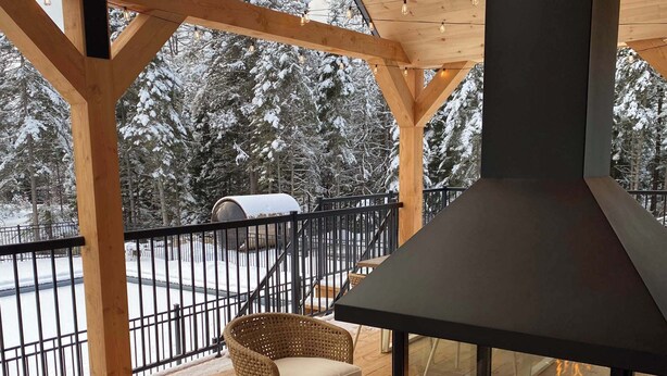 Una terraza techada al aire libre con chimenea de vidrio, sillas de madera y vista a un paisaje nevado.
